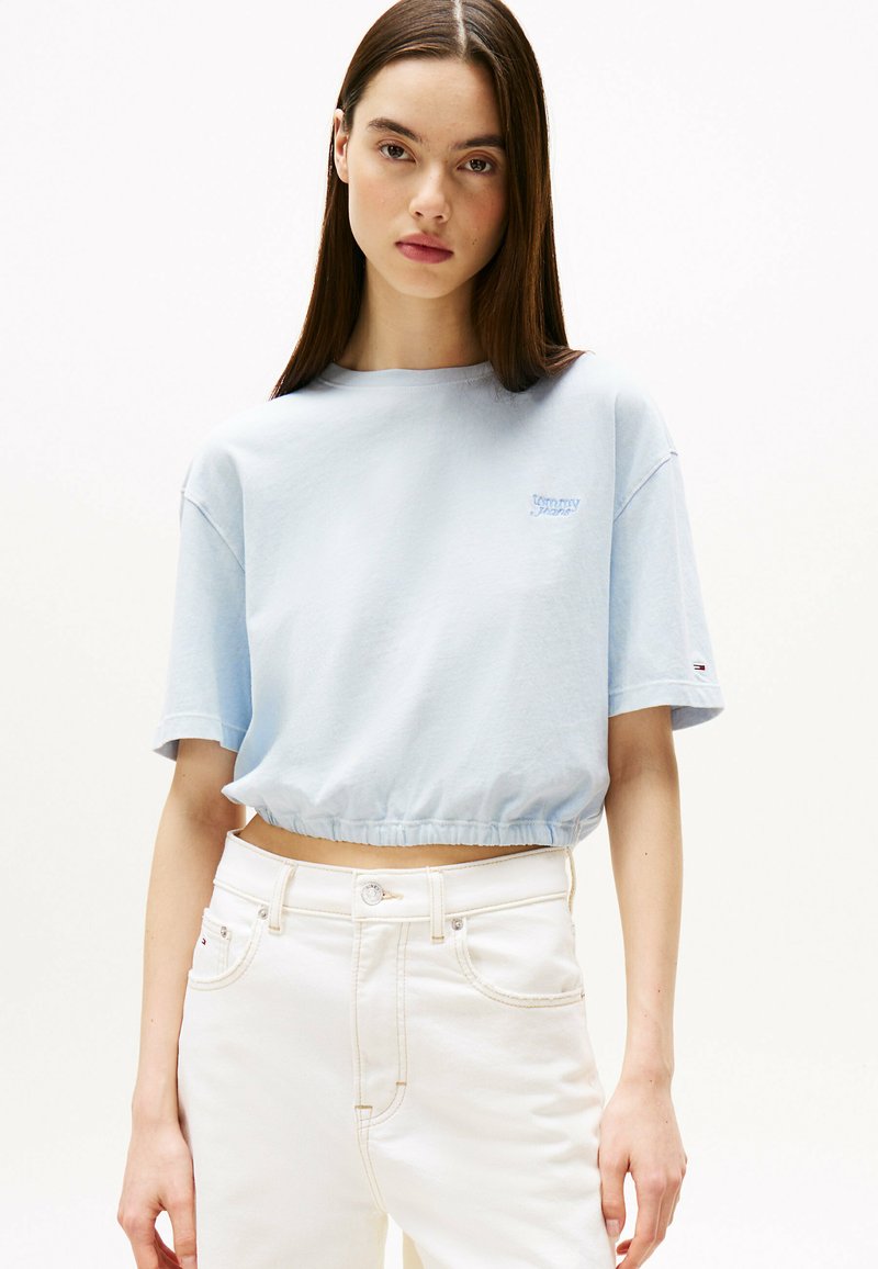 Camiseta corta de color azul claro con mangas cortas, que presenta un discreto logo bordado. Combinada con jeans blancos de cintura alta.