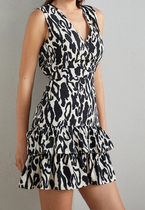 Frau trägt ärmelloses schwarz-weißes Kleid mit Animalprint und gestuftem Rüschenrock vor schlichter Wand.
