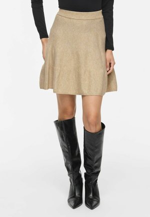 Femme portant une jupe beige longueur genoux associée à des bottes en cuir noires montant jusqu'aux genoux et un haut noir à manches longues, debout sur un fond blanc.