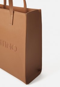 Brun texturerad toteväska med tunna handtag och präglat delvis varumärkesnamn på framsidan, visad mot vit bakgrund.