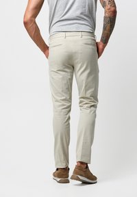 Pantaloni beige dal taglio slim, realizzati in morbido tessuto. Presentano due tasche posteriori con zip. Abbinati a scarpe casual marroni.