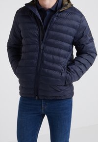 Navy puffer jacket with quilted design, front zip closure, and green hood. Worn over a dark polo and paired with blue jeans.