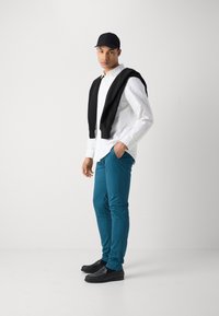 Bela srajca s kratkimi rokavi, črno pulover oblečen čez ramena, teal slim-fit hlače, črni loaferji in črna kapa. Gladke teksture, sproščen stil.