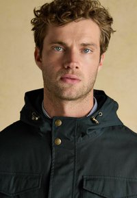 Jachetă impermeabilă verde închis, cu glugă, dotată cu fermoar frontal, două buzunare pe piept, accente din metal auriu și un guler interior cu dungi.