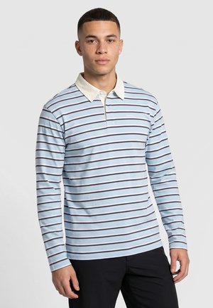 Junger Mann trägt ein langärmeliges, hellblaues Poloshirt mit weißen und schwarzen horizontalen Streifen, steht vor einem schlichten Hintergrund.