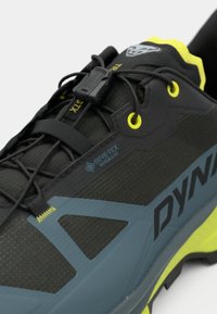 Μαύρο και γκρίζο παπούτσι trail running με επένδυση GORE-TEX. Διαθέτει υφή στην πάνω πλευρά, φωτεινές κίτρινες λεπτομέρειες και δυναμικό σύστημα κορδονιών.