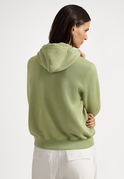 Lysegrønn strikket hettegenser med teksturert overflate, med snøring i hetten og ribbet kant. Brukt med hvite bukser og sett bakfra.