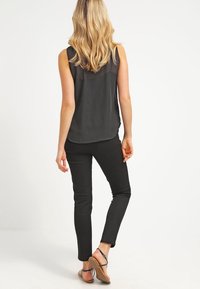 Mujer con cabello rubio vestida con una blusa negra sin mangas con estampado, pantalones negros y sandalias caminando de espaldas sobre un fondo blanco.