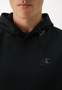 Schwarzer Kapuzenpullover aus weichem Stoff. Verfügt über eine Kordelzugkapuze, ein dezentes Logo auf der Brust und eine glatte Textur mit einem minimalistischen Design.