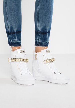 White wedge sneakers with gold chain detail worn with frayed hem blue skinny jeans against a plain light background.