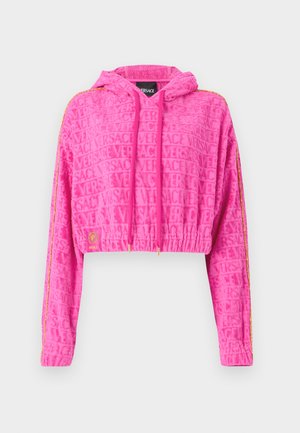 Hoodie cropped di un brillante colore rosa con tessuto testurizzato che presenta il logo "VERSACE" ripetuto. Include un cappuccio, cordini e strisce dorate accentate.