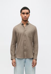 Hnedá košeľa na gombíky s jednou náprsní vreckom, dlhými rukávmi a štandardným golierom, vyrobená zo tkaniny. Kombinovaná so svetlomodrými džínsami.