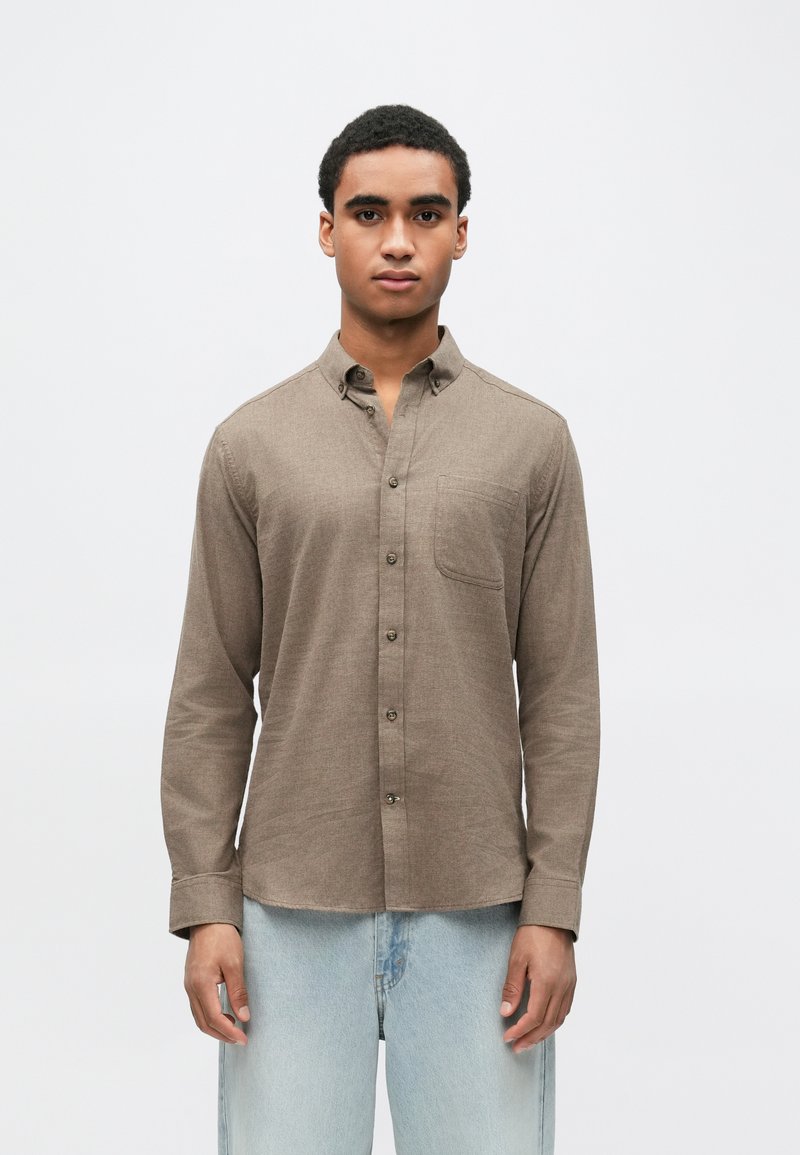 Hnedá košeľa na gombíky s jednou náprsní vreckom, dlhými rukávmi a štandardným golierom, vyrobená zo tkaniny. Kombinovaná so svetlomodrými džínsami.
