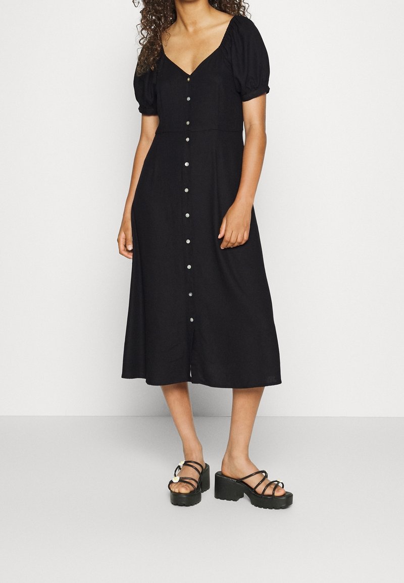 Femme portant une robe midi noire à boutons sur le devant, manches bouffantes et sandales plateformes noires à fines lanières.