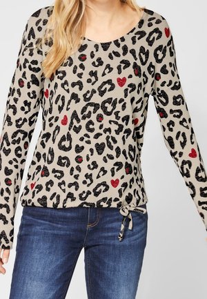 Frau trägt ein beigefarbenes Langarmshirt mit schwarzen Leopardenflecken und roten Herzen, kombiniert mit blauen Jeans und einem Kordelzugsaum.