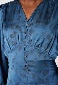 Robe en satin bleu avec motif cachemire foncé, boutons recouverts de tissu au centre, taille froncée et décolleté en V.