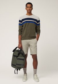 Maglione a righe verdi e grigie, pantaloni corti beige, con uno zaino verde scuro dotato di molte tasche e cerniere, abbinato a sneaker chiare.
