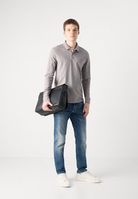 Black messenger bag with a sleek design, shown held by a person wearing a light gray long-sleeve polo and blue jeans. White sneakers visible.