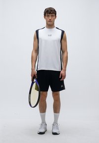 Camisola branca desportiva sem mangas para homem com detalhes em azul-marinho, combinada com calções pretos. Ele segura uma raquete de ténis e usa meias e sapatilhas desportivas brancas.