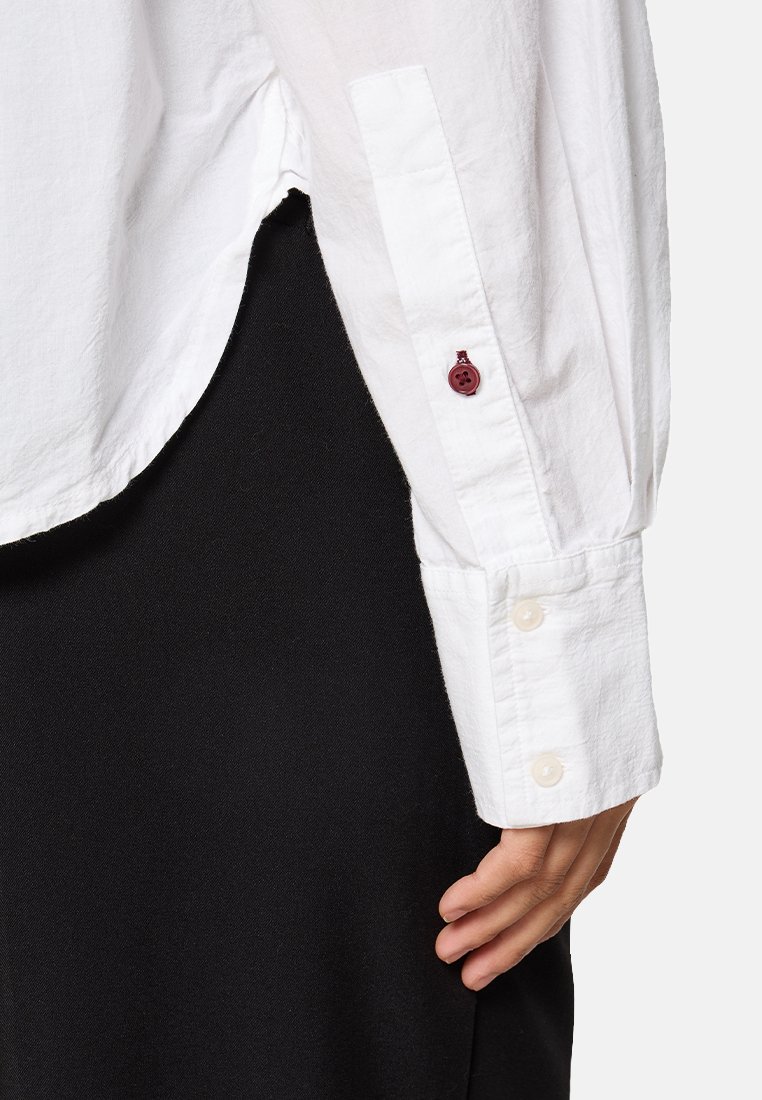 Witte langarmshirt van een licht gestructureerde stof, met een dubbele manchetknoop in crème en een kastanjebruine knoopaccent bij de mouwkop.