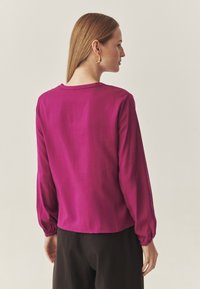 Blusa fucsia a maniche lunghe con scollo rotondo e polsini elasticizzati, realizzata in tessuto leggero, con una consistenza liscia e una vestibilità comoda.