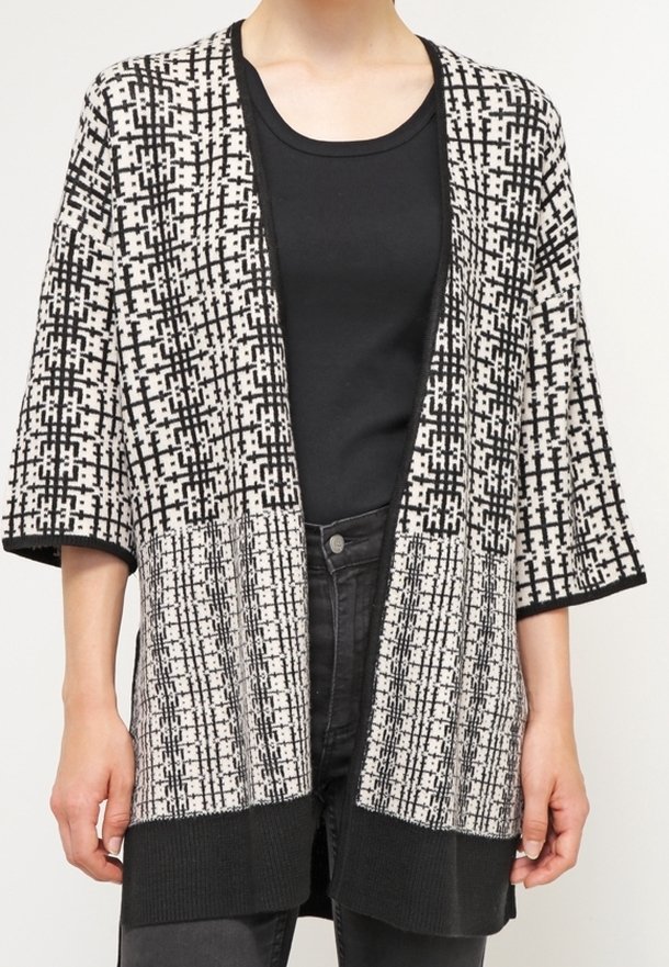 Cardigan en tricot avec un motif géométrique noir et blanc, devant ouvert et manches larges. Doté d'un ourlet noir uni. Porté sur un haut noir.