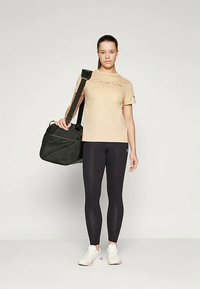 Beige t-shirt με το λογότυπο "Champion", μαύρες κολάν, κρατώντας μια μαύρη τσάντα γυμναστικής, λευκά sneakers με μωβ λεπτομέρειες, απαλές υφές.