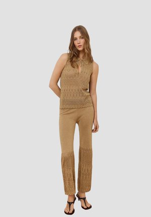Vrouw draagt een beige mouwloos gebreid topje met een strik aan de hals en bijpassende gebreide broek met een patroon aan de onderkant, staand tegen een egale achtergrond.