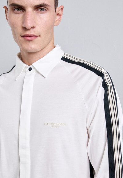 Vit polo med knäppt krage, med marinblå ränder längs axlarna och en broderad logotyp på bröstet.