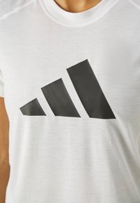Hvid atletisk t-shirt med korte ærmer, der har et stort sort geometrisk logo på fronten. Fremstillet af letvægtsstof med en glat tekstur.