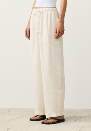 Persoon draagt losse, crèmekleurige broek met elastische tailleband en trekkoord, gecombineerd met zwarte slippers, staand op een stenen vloer.
