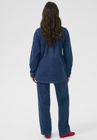 Hosszú ujjú, egyenes szabású, kerekített hemű farmering, amelyet hozzá illő bőszárú farmernadrággal és vörös cipővel viselnek. Texturált anyag.