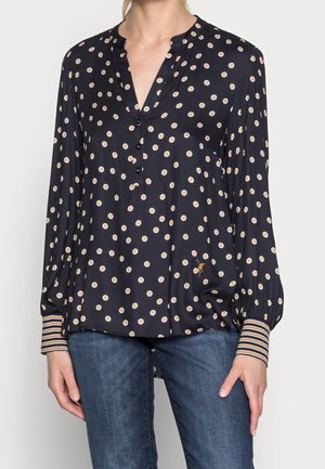 Kobieta ubrana w granatową bluzkę z długim rękawem w beżowe i białe kropki oraz mankiety w paski, do tego niebieskie dżinsy.