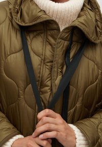 Olivengrøn quiltet dunjakke med hætte og sorte snore, båret over en cremefarvet ribbet rullekravesweater.