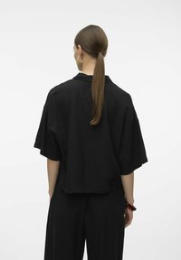 Top corto nero a maniche corte realizzato in tessuto testurizzato, caratterizzato da una vestibilità comoda e colletto aperto. La vista dal retro mostra un'acconciatura con coda di cavallo lunga fino alle spalle.