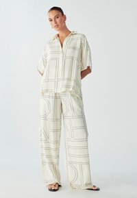 Conjunto ligero de color crema, compuesto por una blusa de mangas cortas oversized y pantalones de pierna ancha, que presentan contornos y líneas geométricas en negro.