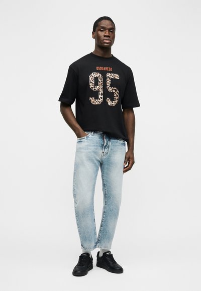 Άνδρας που στέκεται με το χέρι στην τσέπη, φοράει μαύρο μπλουζάκι "DSQUARED2" με τύπωμα λεοπάρδαλης και τον αριθμό "95", ανοιχτόχρωμο τζιν και μαύρα αθλητικά παπούτσια σε λευκό φόντο.