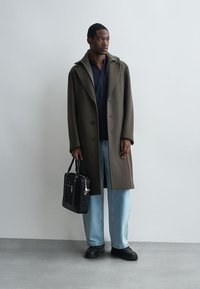 Ελιά πράσινο μάλλινο παλτό με μεγάλες παρενθέσεις, κλείσιμο με κουμπί, φορεμένο πάνω από ένα σκούρο πουκάμισο και ανοιχτό μπλε τζιν; κρατώντας μια μαύρη, δομημένη τσάντα.