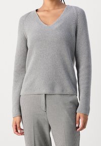 Femme portant un pull V en maille côtelée gris clair et un pantalon à carreaux noir et blanc, debout avec les mains détendues sur les côtés.
