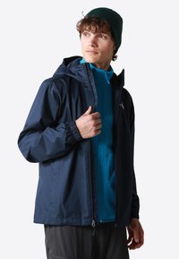 Chaqueta impermeable azul marino con capucha, puños elásticos y cremallera frontal. Llevada sobre una chaqueta de media capa texturizada en color agua.