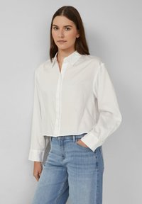 Joven con cabello castaño liso, vistiendo una camisa blanca de botones y jeans de un azul claro, de pie con una mano en el bolsillo.