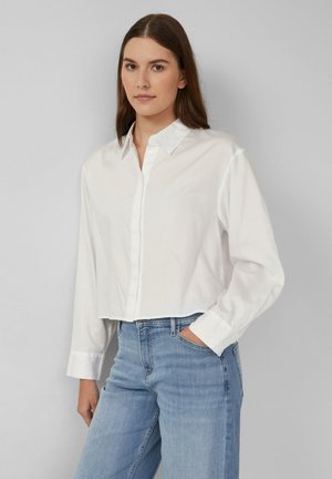 Giovane donna con capelli castani lisci, che indossa una camicia bianca con bottoni e jeans azzurri chiari, in piedi con una mano in tasca.