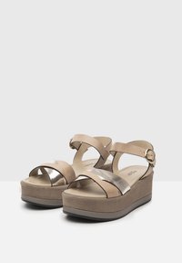 Beige klínové sandály s širokými křížovými pásky, metalickými akcenty a nastavovacími popruhy na kotníku. Měkký, texturovaný svršek s polstrovanou stélkou.