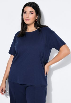 Frau mit langen dunklen Haaren, die ein marineblaues kurzärmeliges Hemd und Hose trägt und mit einer Hand auf der Hüfte vor weißem Hintergrund steht.