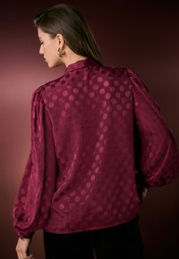 Blusa bordeaux a pois con maniche a sbuffo, texture liscia e collo con bottoni. Accentuata da una sottile lucentezza nel tessuto.