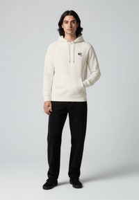 Light beige hoodie with a front pocket and drawstring, featuring a small logo on the chest, paired with black pants and black shoes.
