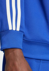 Blauer Sportpulloverärmel mit drei weißen Streifen. Material ist glatt mit einem taillierten Bund und strukturiertem Detail auf dem Stoff.