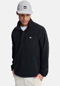 Sudadera de forro polar negra con cuello alto, tapeta de botones y bolsillo frontal; cuenta con un pequeño logo y un acabado suave y texturado.
