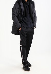 Chaqueta acolchada negra con capucha, suéter oscuro texturizado, pantalones negros con el logo FILA en rojo y blanco, y zapatos negros con suelas gruesas.