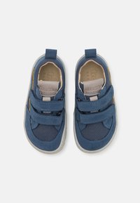 Blaue Stoffschuhe mit Netzpaneelen, doppelten Klettverschlüssen, einer hellgrauen Sohle und einem kleinen gelben Akzent. Weiche Innenauskleidung.